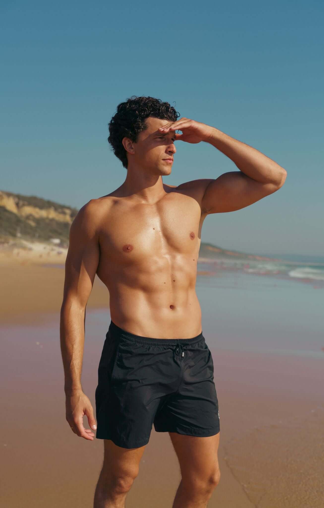 Man in black swim shorts standing on the beach, hand shielding eyes from sun.