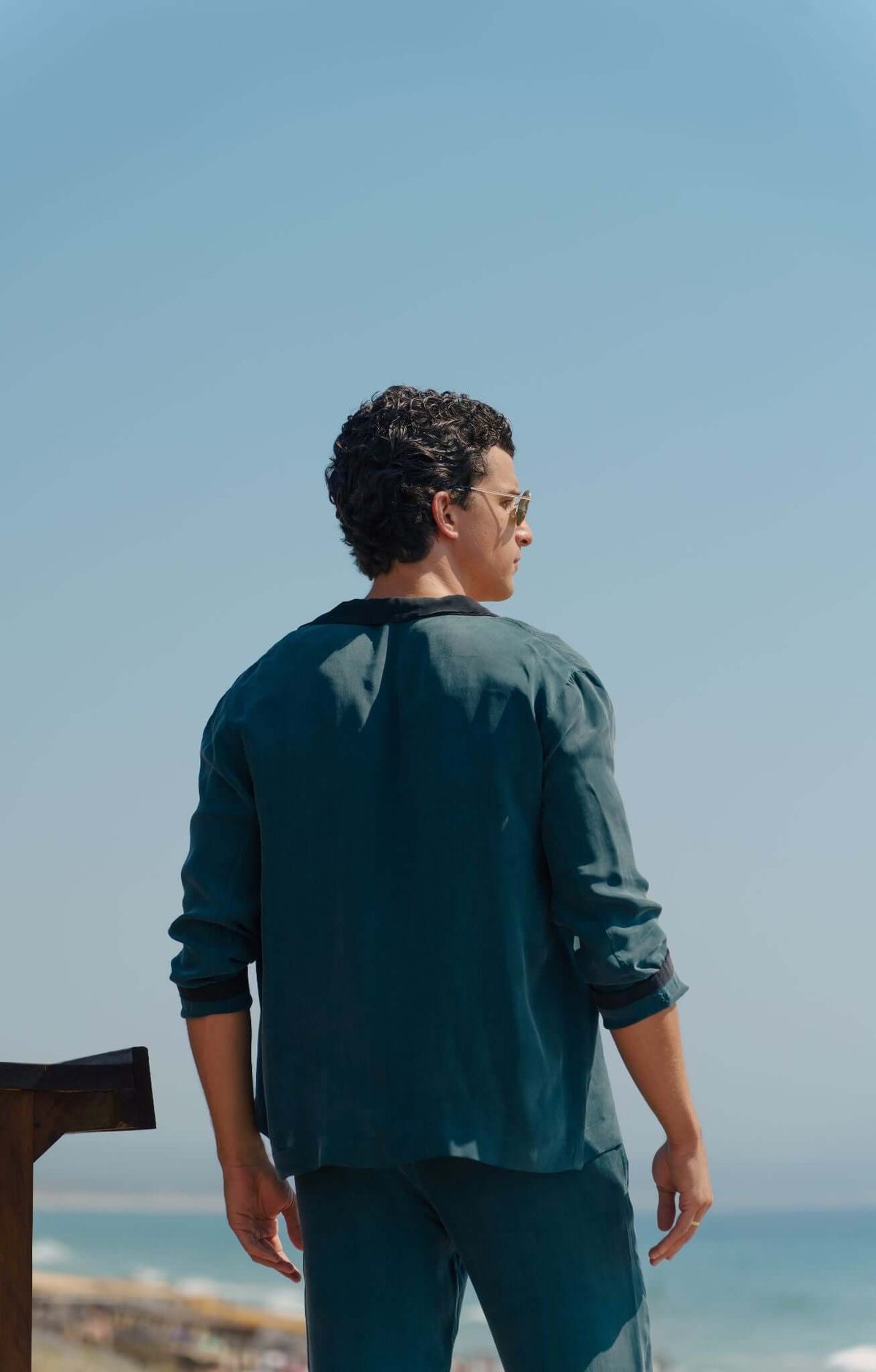 Man wearing a dark green Pyjama Set, viewed from the back, against a clear blue sky and ocean.