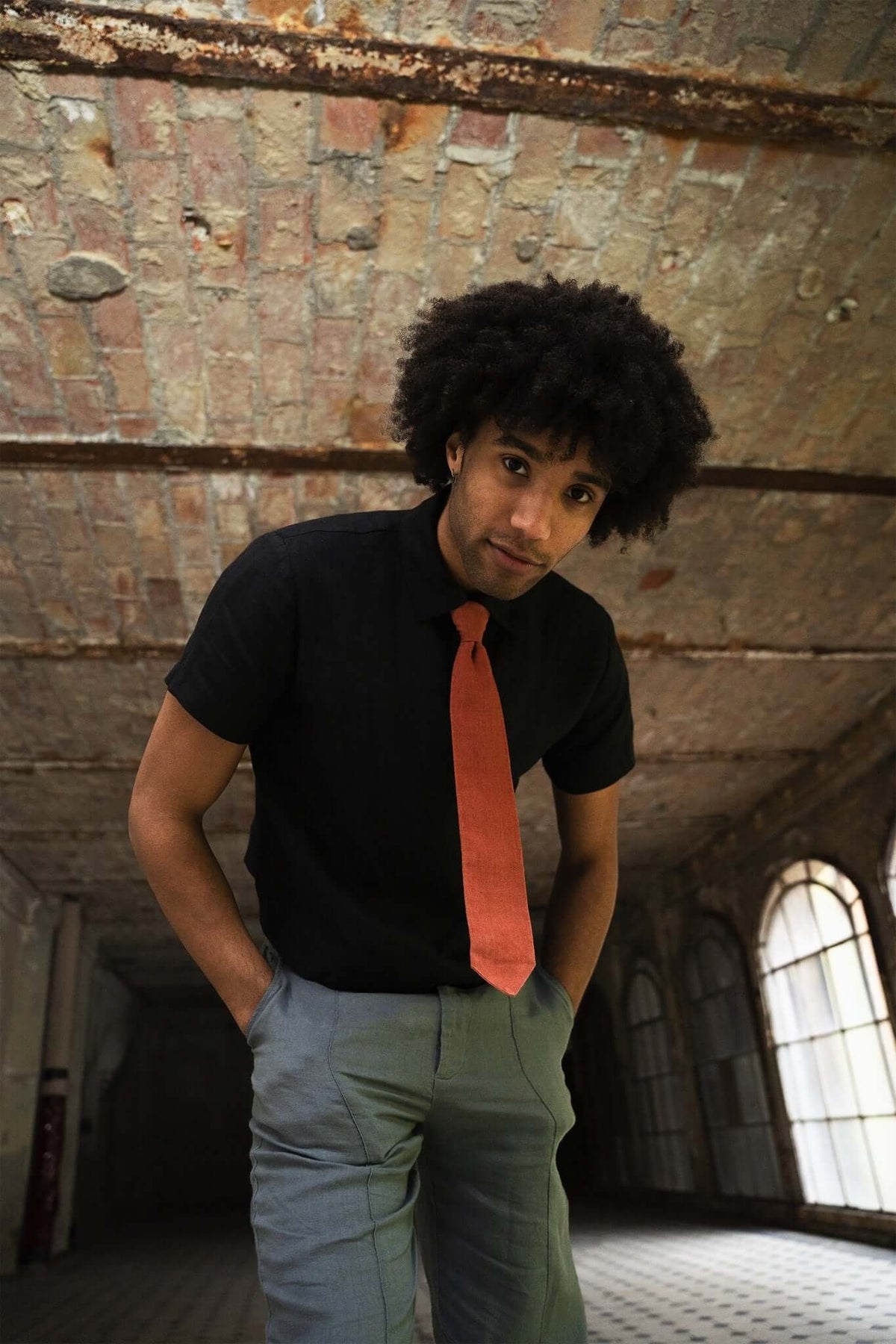 Man with afro hairstyle in a black shirt and orange tie, standing in a rustic industrial space.