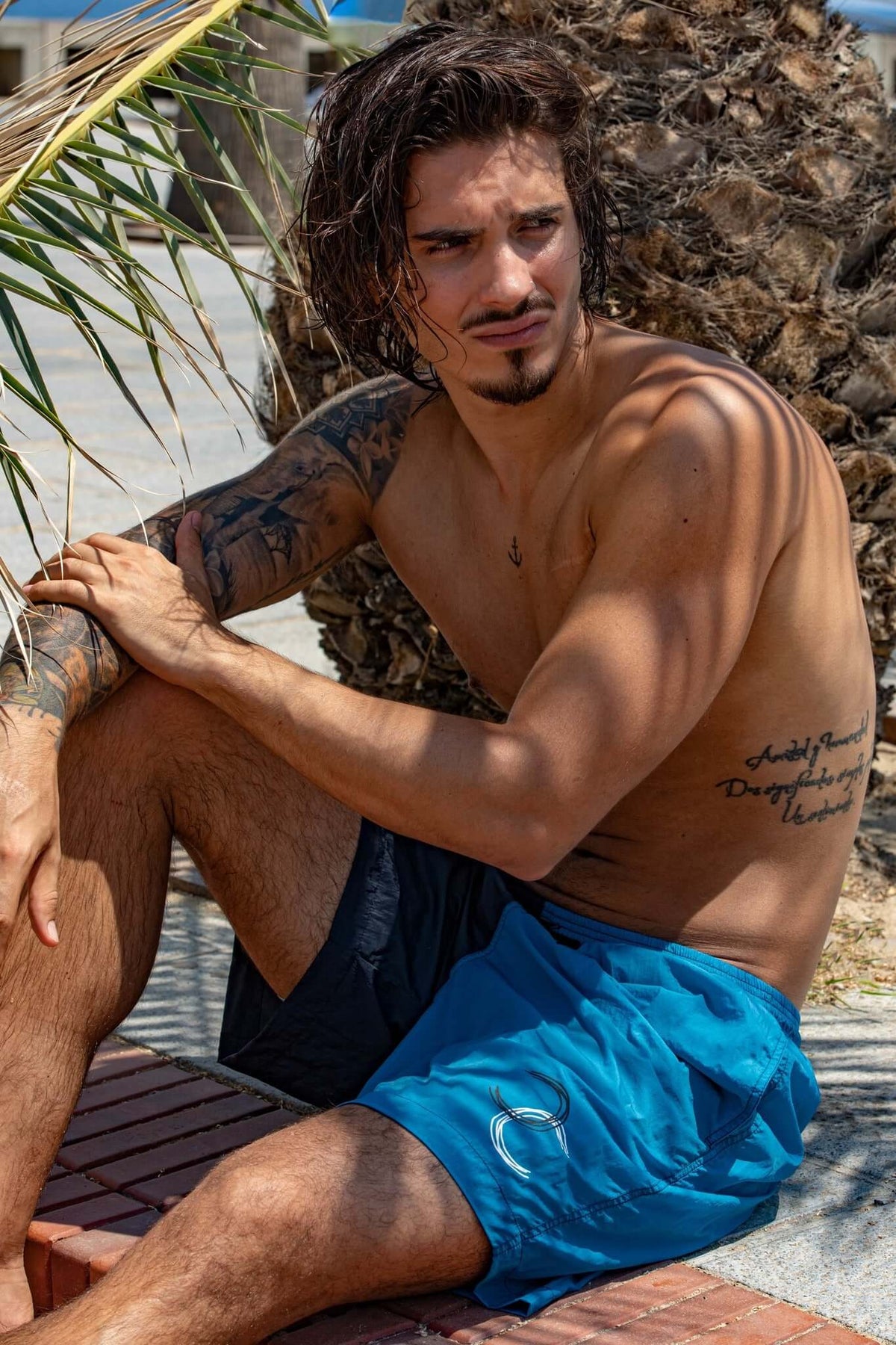 Man with tattoos sitting casually against a palm tree, wearing blue swim shorts in a sunny outdoor setting.