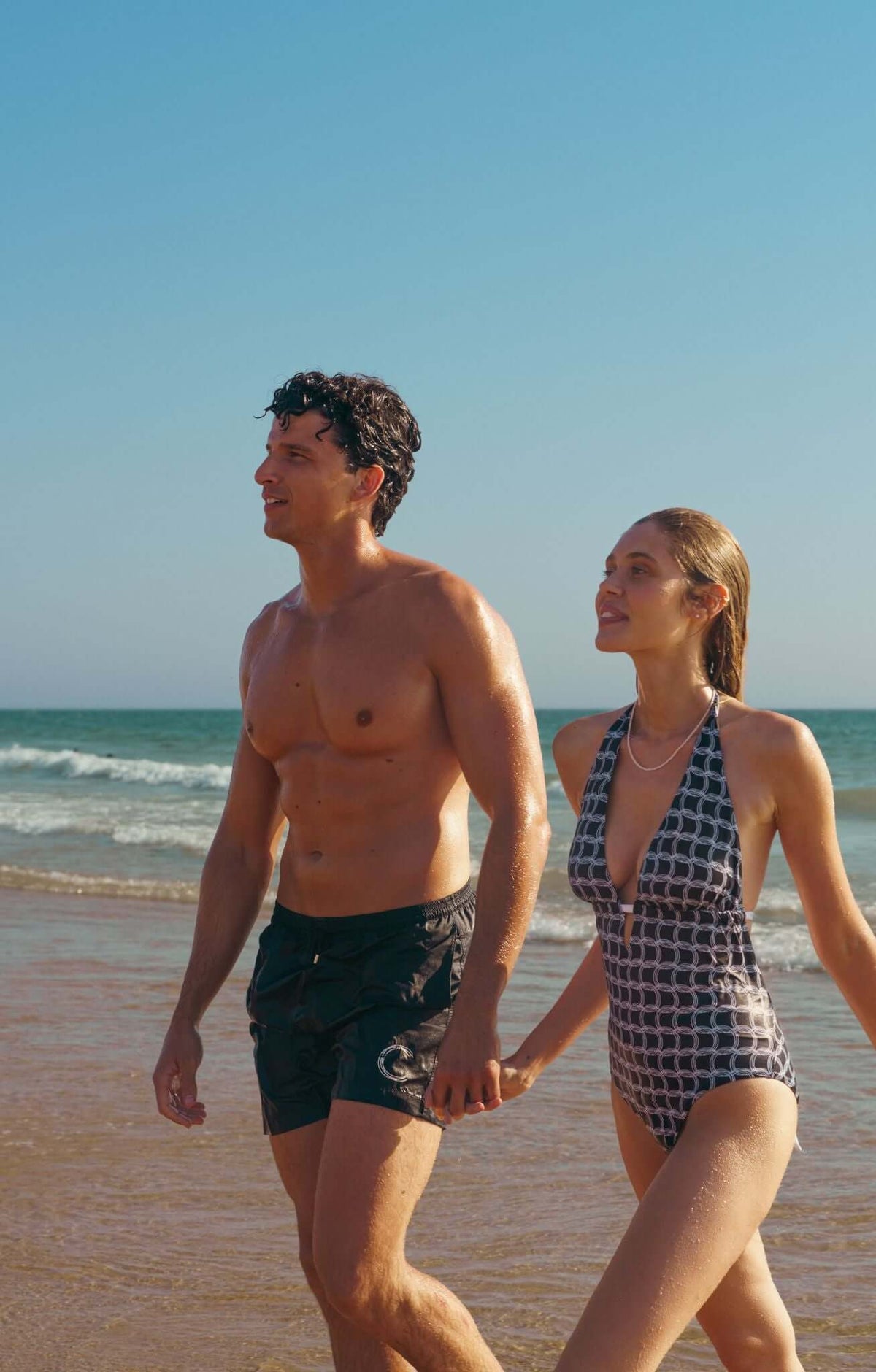 Man and woman walking hand in hand on a beach, ocean waves in the background, summer vibes.