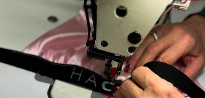 Hands sewing a black label on pink fabric using an industrial sewing machine.
