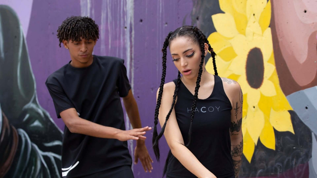 Two dancers in HACOY apparel performing in front of a vibrant mural with sunflowers.