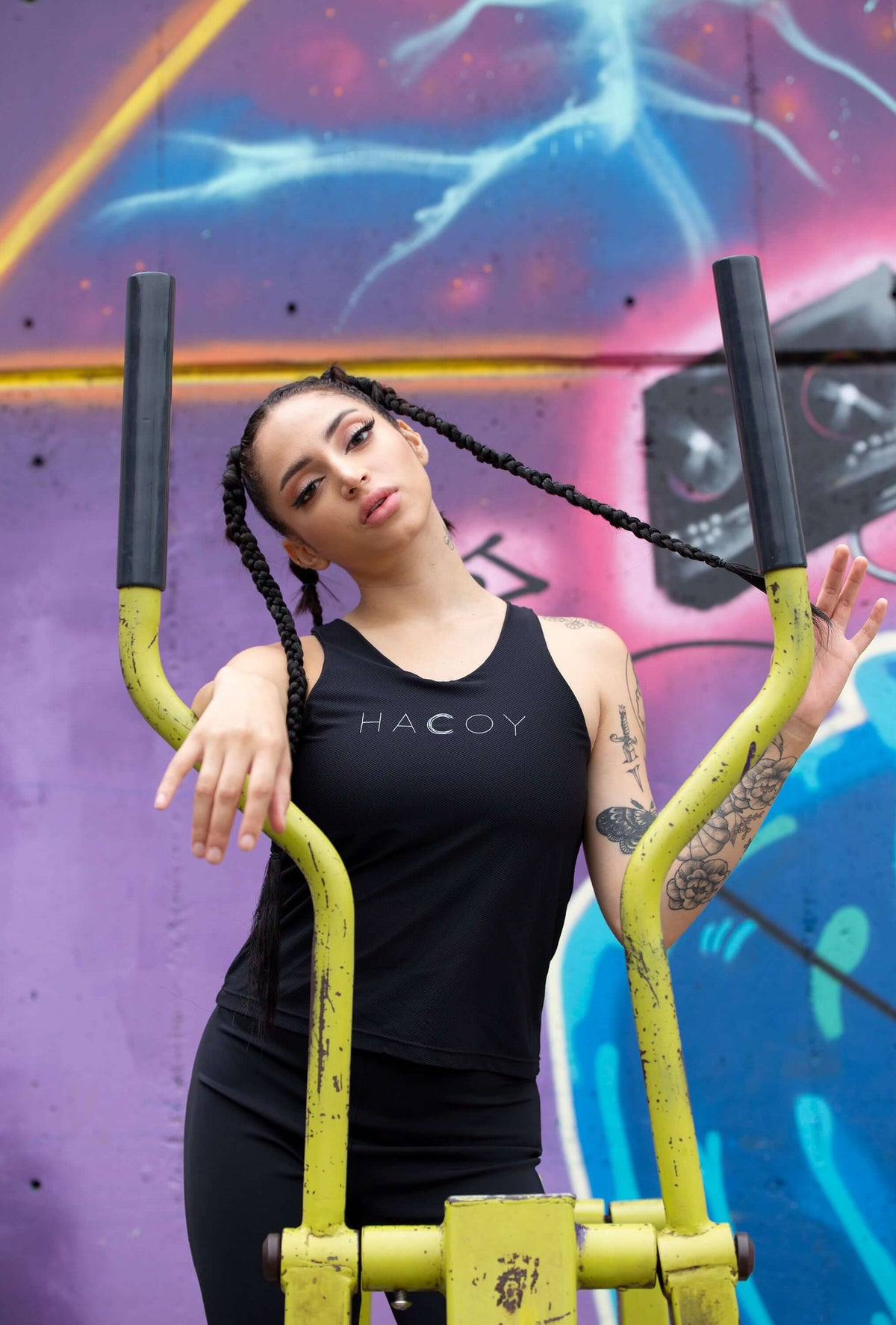 Model in HACOY tank top poses confidently by gym equipment against vibrant graffiti backdrop.