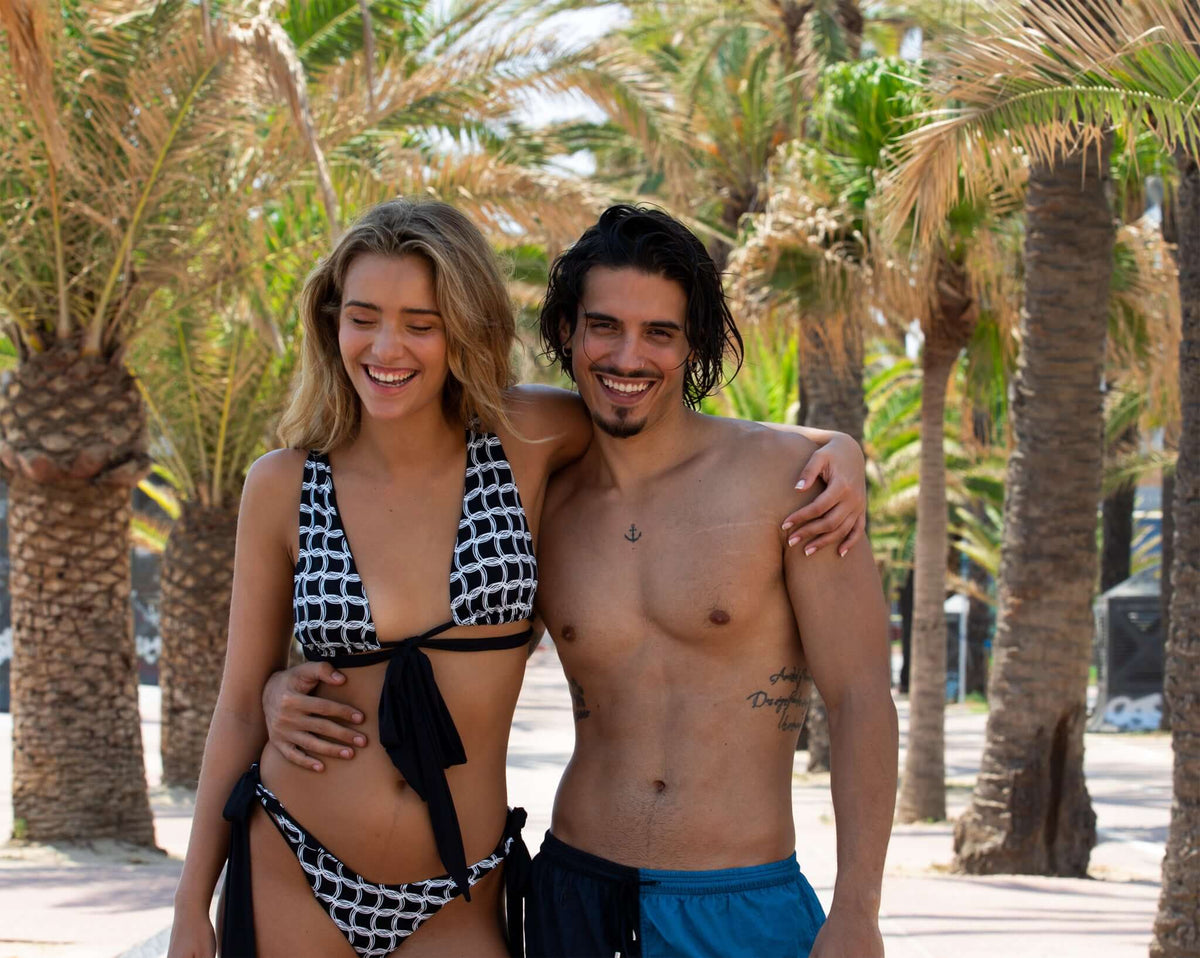Couple in swimwear smiling together on a palm tree-lined beach during sunny weather.