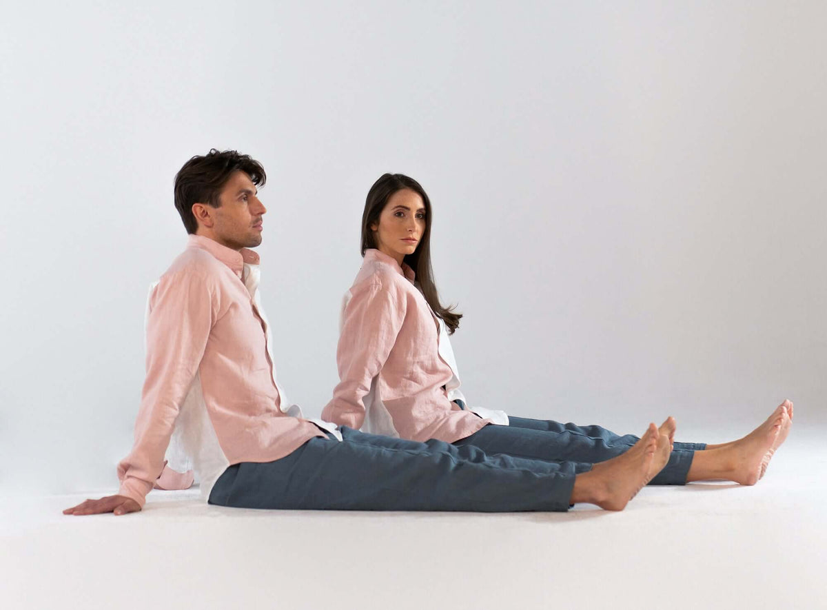 A man and woman sitting on the floor, both wearing casual pink shirts and relaxed blue pants, against a neutral background.